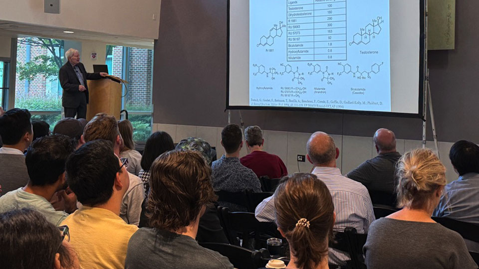 Audience watching a presenter give a scientific lecture with chemical structures and data displayed on a large projection screen.