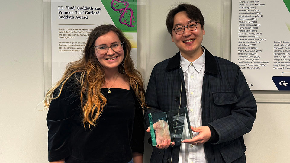 A woman with long brown hair wearing a black blouse smiles for a photo next to a man with black hair wearing a white shirt and black jacket. The main holds a glass award. 