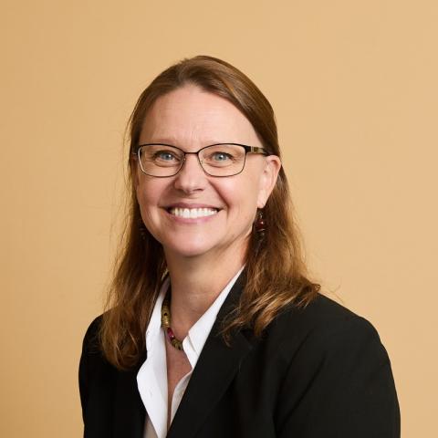 A woman with shoulder-length brown hair and glasses smiles for a portrait in front of a yellow backdrop.